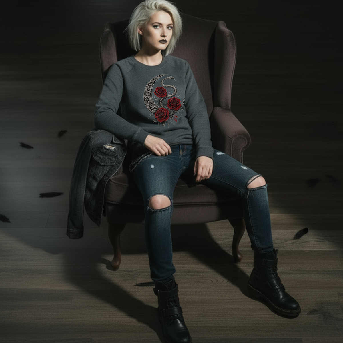 Woman sitting in a dark room wearing a gray sweatshirt with red floral design, ripped jeans, and black boots.